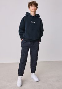 Mørkeblå hættetrøje med teksten "BAKER" og en kengurulomme. Kombineret med mørke cargobukser og hvide sneakers. Enkel, funktionel design.