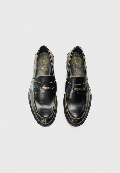 Chaussures en cuir noir avec une finition lisse et des coutures beiges, comportant une sangle en métal sur le dessus. Semelles intérieures à motif floral visibles.