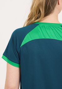 Mørk teal t-shirt med grønne detaljer og rund halsudskæring. Har korte ærmer og en subtil tekstur. Ingen synlige mønstre eller hardware-detaljer.