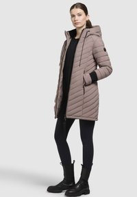 Kvinde iført en lang mauve quilted frakke med hætte, sorte leggings og sorte ankelstøvler, stående med den ene hånd i lommen.