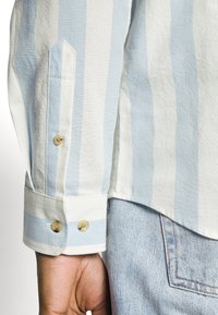 Una camicia a righe blu chiaro e bianco con dettagli sui polsini abbottonati. Un tessuto morbido con una vestibilità rilassata e cuciture curate, abbinata a denim.
