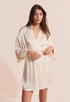 Jeune femme aux longs cheveux bruns portant un court peignoir en satin crème, ajustant la ceinture sur un fond beige clair.