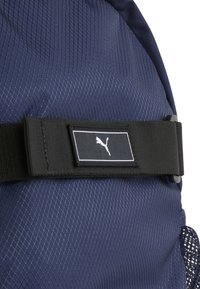 Mochila navy con un tejido texturizado, que cuenta con una correa negra con una etiqueta de logo blanca y un bolsillo lateral de malla. Patrón de diamante visible.