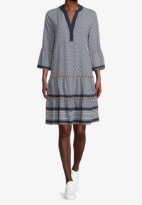 Femme portant une robe à motifs arrivant aux genoux avec des manches évasées et des baskets blanches, se tenant devant un fond blanc uni.