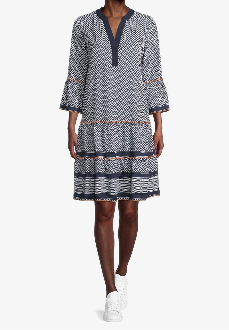 Femme portant une robe à motifs arrivant aux genoux avec des manches évasées et des baskets blanches, se tenant devant un fond blanc uni.