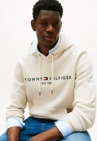 Felpa beige con logo e data di fondazione, dotata di lacci e di una consistenza morbida, indossata sopra una camicia a righe blu.