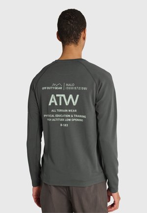 Mann trägt dunkelgraues Langarmshirt mit Off-Duty-Ausrüstung und ATW-Schriftzug auf dem Rücken, kombiniert mit dunkler Hose, steht vor weißem Hintergrund.