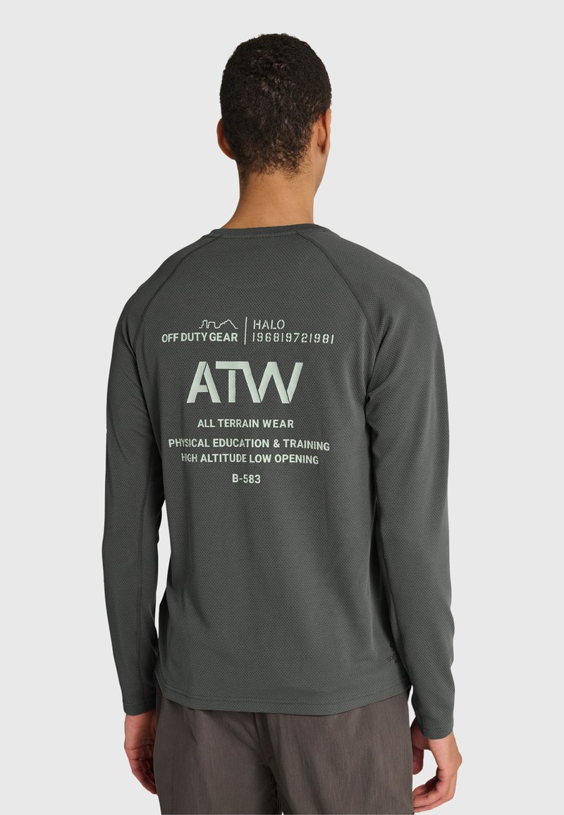 Mann trägt dunkelgraues Langarmshirt mit Off-Duty-Ausrüstung und ATW-Schriftzug auf dem Rücken, kombiniert mit dunkler Hose, steht vor weißem Hintergrund.