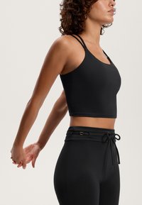 Čierny skrátený top bez rukávov má výstrih do oblúka a dvojité ramienka. Čierne legíny s vysokým pásom majú nastaviteľný pás a ozdobné kovové detaily.