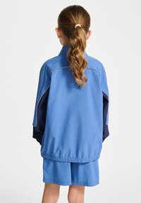 Blå idrottsjacka med en texturerad yta och marinblå av detaljer, tillsammans med matchande blå shorts. Har elastiska manschetter och nederkant.