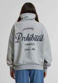 Sudadera gris de gran tamaño con cuello alto, que presenta el texto azul marino "Prendas Prohibidas" y elementos decorativos en la parte posterior.