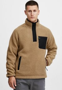 Sudadera de forro polar beige con un bolsillo en el pecho con cremallera negra y bolsillos laterales, con un cuello alto y tejido texturizado.