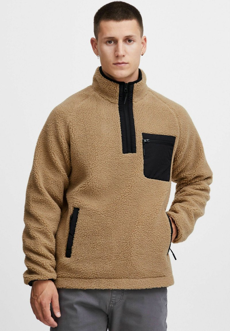 Sudadera de forro polar beige con un bolsillo en el pecho con cremallera negra y bolsillos laterales, con un cuello alto y tejido texturizado.