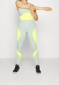 Top corto atletico grigio chiaro con accenti giallo neon abbinato a leggings coordinati con un simile blocco di colori audaci e scarpe sportive nere.