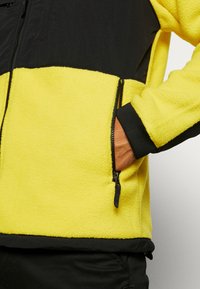 Fleeceová bunda s jasně žlutou spodní částí a černou horní částí. Má kapsu na zip a kontrastní černé manžety. Jemná struktura.
