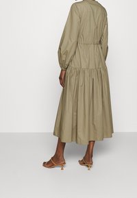 Donna che indossa un lungo e ampio abito verde oliva con vita arricciata e maniche, abbinato a sandali con tacco kitten aperti in punta color beige, di spalle.