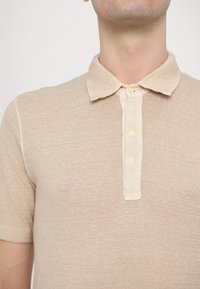 Polo bej realizat dintr-un material texturat, având o deschidere cu nasturi cu patru nasturi crem și un guler moale, ușor ridicat.