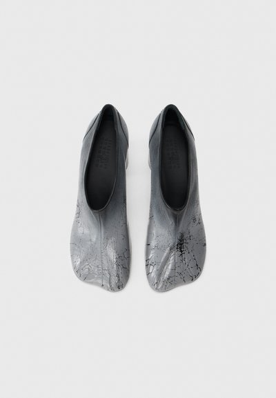 Ζευγάρι μεταλλικών ασημένιων slip-on παπουτσιών με μαύρες ραγισμένες λεπτομέρειες, στρογγυλεμένη τετράγωνη μύτη και χαμηλή κοπή στο άνοιγμα σε λευκό φόντο.