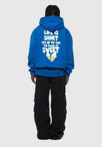 Blå hoodie med stor vit text som säger "LIVET ÄR KORT DET ÄR UPP TILL DIG ATT GÖRA DET SÖTT" och en glassgrafik på baksidan. Svarta cargo-byxor.