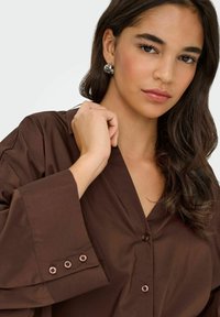 Femme aux cheveux foncés portant une chemise marron boutonnée, des boucles d'oreilles argentées à motifs, tenant le col d'une main sur un fond blanc.