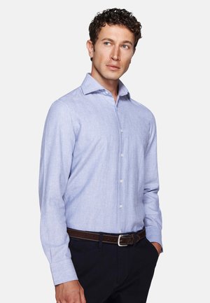 Man with curly hair wearing a light blue long-sleeve button-up shirt, dark pants, and a brown belt, looking to his left with hands in pockets.