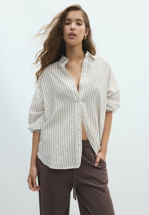 Jeune femme aux cheveux ondulés lâchés porte une chemise beige à rayures blanches boutonnée avec manches retroussées et un pantalon marron foncé, posant la main dans la poche.