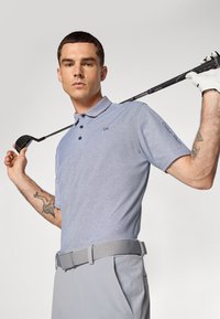 Moški v svetlo modri polo majici in sivih hlačah drži golf palico za rameni, pri čemer pokaže tetovaže na rokah in nosi belo golf rokavico na desni roki.