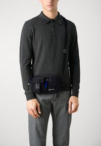 Černá nylonová crossbody taška s několika zipovými compartmenty, modrým zipem a nastavitelným popruhem, nošená přes tmavý svetr a šedé kalhoty.