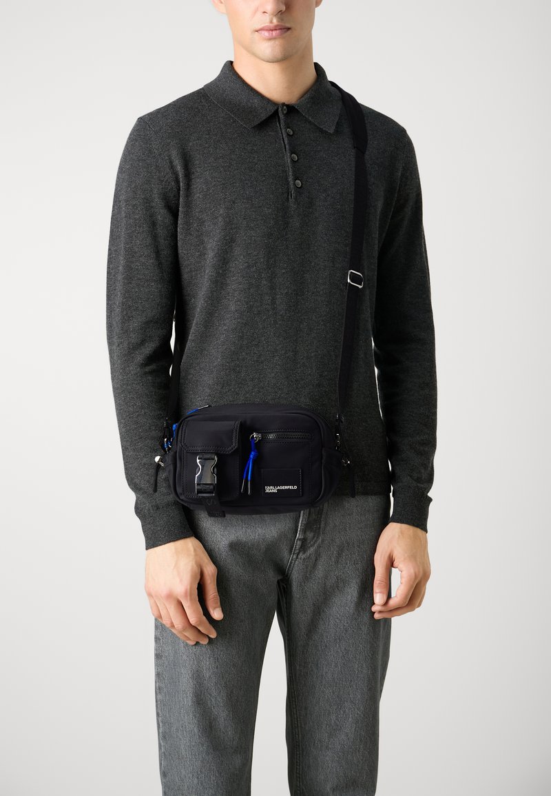 Černá nylonová crossbody taška s několika zipovými compartmenty, modrým zipem a nastavitelným popruhem, nošená přes tmavý svetr a šedé kalhoty.