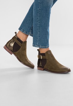 Person iført blå jeans og olivengrønne ankelstøvler med guldbuckle og metallisk hældetalje på en hvid gulv.