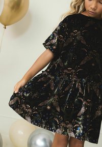 Melnas samta kleita ar īsām piedurknēm. Iezīmēta ar krāsainu ziedu rakstu zaļā, zeltainā un ziliem toņos. Saplacināta svārku daļa ar salocītu jostas daļu.