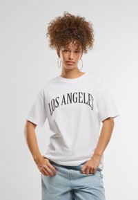 T-shirt en coton blanc avec une coupe oversize, arborant "LOS ANGELES" en lettres noires en gras. Manches courtes, design décontracté. Jean baggy bleu clair.