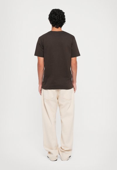 Homme de dos portant un t-shirt à manches courtes marron foncé et un pantalon beige ample, sur un fond clair uni.