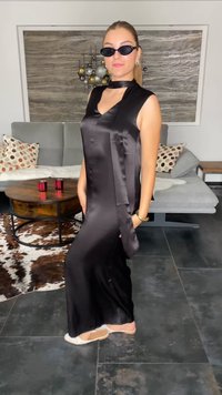 Vestido maxi de satén negro con un cuello alto, diseño sin mangas y detalle de lazo lateral. Presenta una textura suave y se lleva con sandalias blancas.