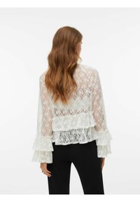 Hvit bluse i blonder med rysjedetaljer, gjennomsiktig tekstur, vide ermer og lagdelt design, kombinert med svarte bukser.