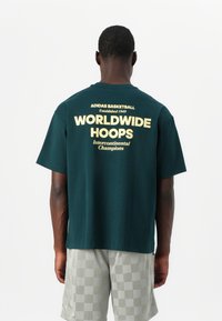 Donkergroen katoenen t-shirt met korte mouwen, met daarachter gele bedrukte tekst: "ADIDAS BASKETBALL," "WORLDWIDE HOOPS," en "Intercontinental Champions."