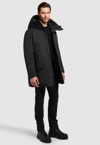 Muž v černém zatepleném kabátu s kapucí, černém svetru, černých džínách a černých botách, stojící na jednobarevném šedém pozadí.