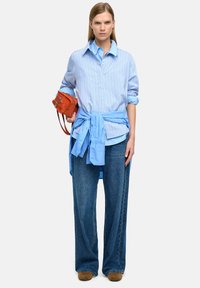 Kobieta stoi, ubrana w niebieską koszulę w paski, niebieską kurtkę związaną w talii, szerokie niebieskie dżinsy, brązowe buty i trzyma małą pomarańczową torebkę.