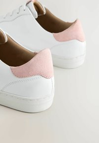 Baskets blanches avec des patchs de talon en daim rose clair, coutures visibles, doublure intérieure beige et semelles extérieures en caoutchouc blanc texturé, posées sur une surface claire.