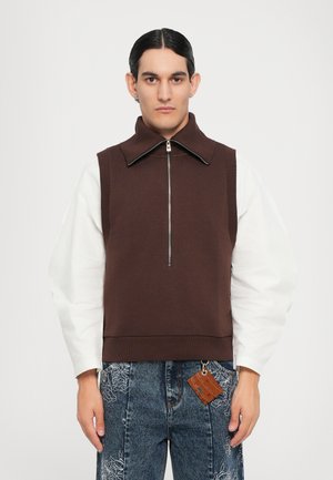 Brauner ärmelloser Sweatshirt mit hohem Kragen und Reißverschluss. Kombiniert mit weißen Langarmshirts und Jeans mit floralen Stickereien.