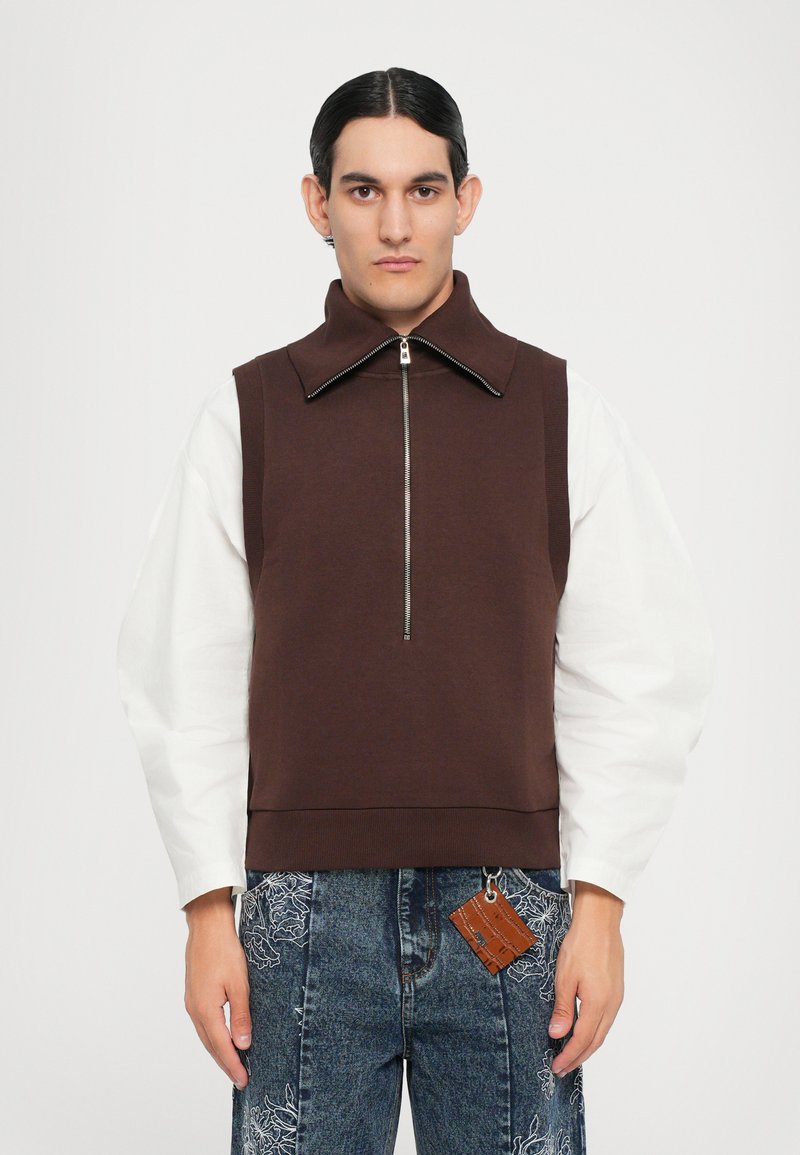 Brauner ärmelloser Sweatshirt mit hohem Kragen und Reißverschluss. Kombiniert mit weißen Langarmshirts und Jeans mit floralen Stickereien.