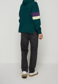 Sudadera con capucha en azul verdoso con rayas moradas y amarillas; combinada con jeans gris oscuro y zapatillas blancas con suela marrón.