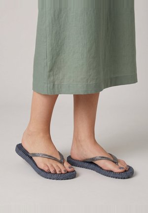 Im Schatten stehende Füße, die graue Zehensandalen mit geflochtenen Sohlen tragen, auf einem neutralen Boden, mit einem grünen Rocksaum sichtbar über den Knöcheln.