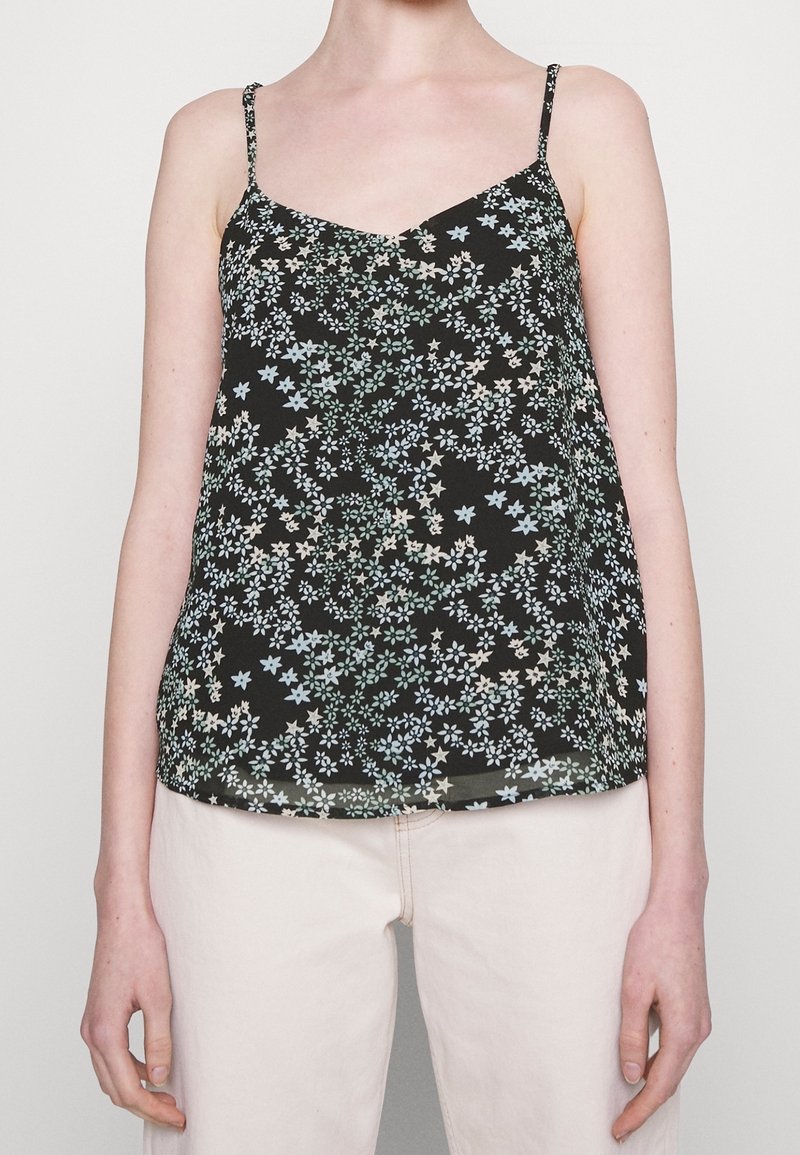 Personne portant un haut noir à fines bretelles à motif floral et un pantalon clair sur un fond uni.