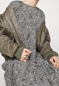 Oliivinvihreä pommitakki, jossa on ribbireunat, kerrostettuna beigen mekon päällä, jossa on musta abstrakti kuvio ja röyhelöhelma.