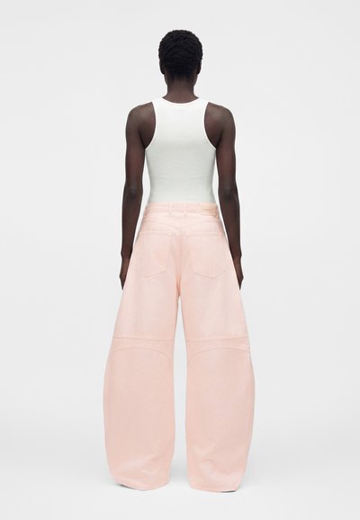 Άτομο που φαίνεται από πίσω, φοράει λευκό αμάνικο τοπ και ανοιχτό ροζ φαρδιά παντελόνια και στέκεται μπροστά σε απλό φόντο.