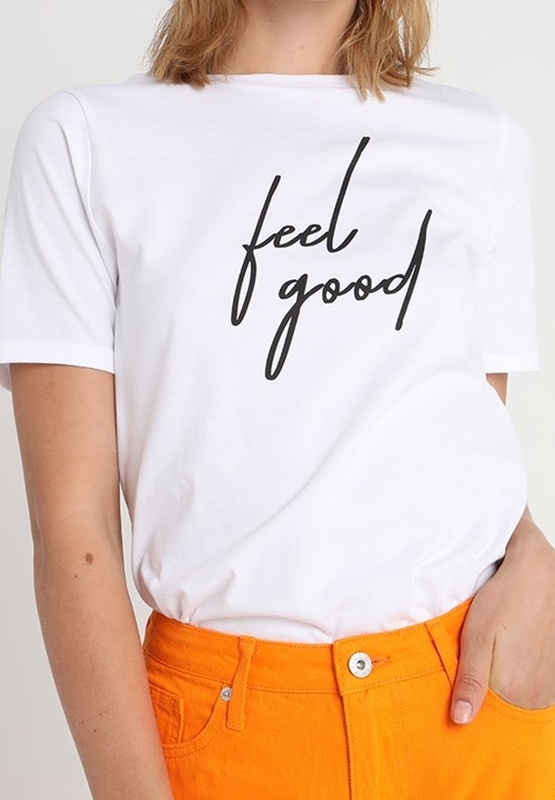 T-shirt blanc avec texte noir "feel good", porté avec un pantalon taille haute orange vif, main reposant dans la poche.