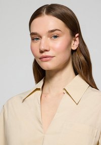 Camicia beige con colletto e scollatura a V profonda, dotata di taschino sul petto e logo discreto. Tessuto liscio con una leggera lucentezza.