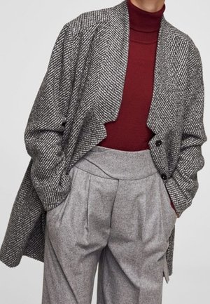 Femme portant un manteau gris à chevrons par-dessus un col roulé bordeaux et un pantalon gris plissé taille haute, les mains dans les poches.