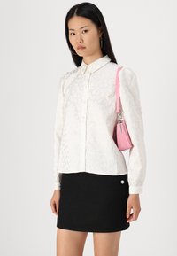 Hvit teksturert bluse med krage, kombinert med et svart miniskjørt med en sølvknappdetalj, og en rosa håndveske.